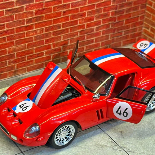 A red die-cast model sports car with blue and white racing stripes, open doors, and race number decals, photographed indoors against a brick wall and tiled floor.